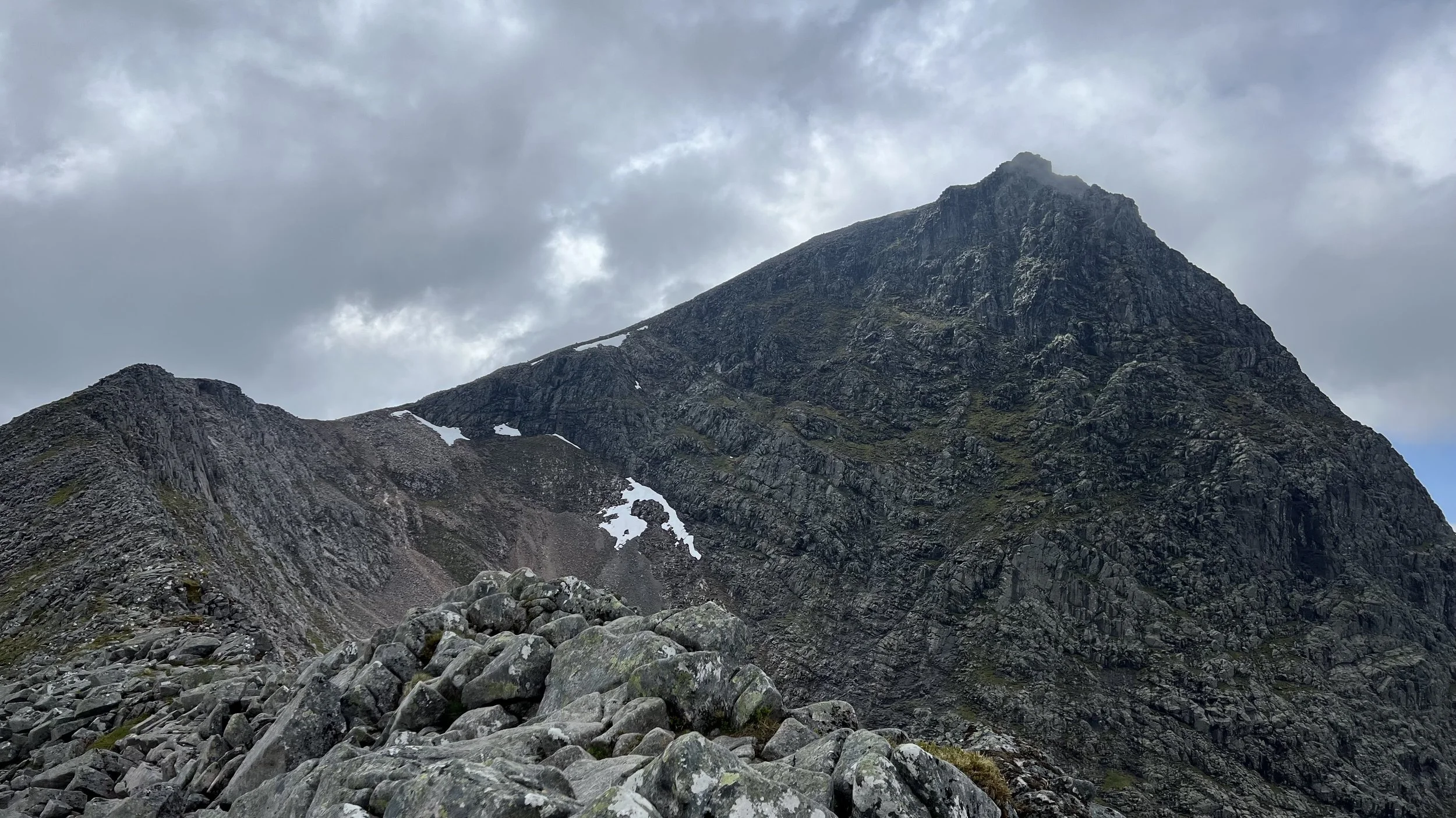 A rugged mountain landscape with a steep peak, patches of snow, and a cloudy sky.