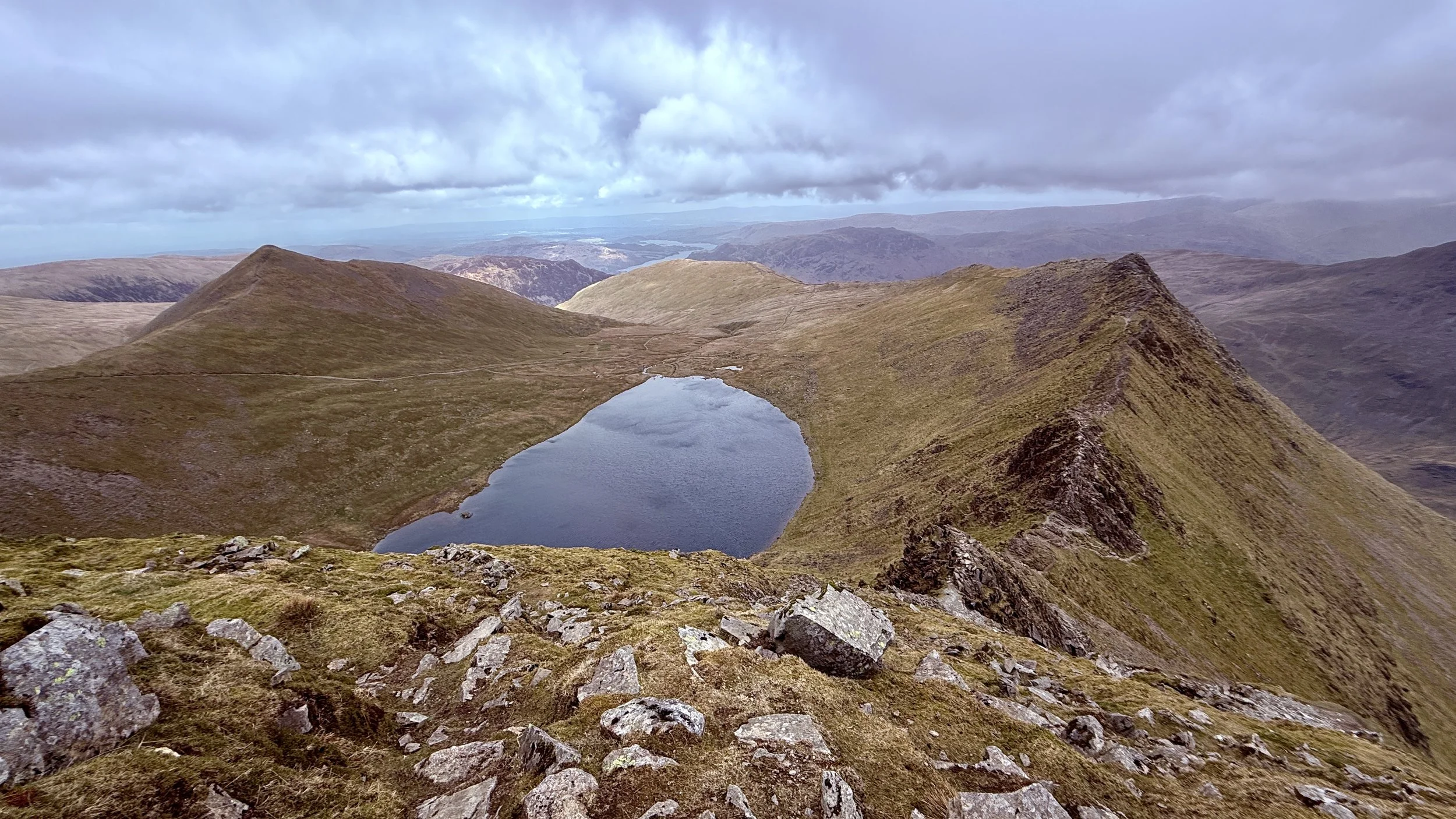 A mountain landscape with a small lake in a basin surrounded by grassy, rocky slopes under a cloudy sky.