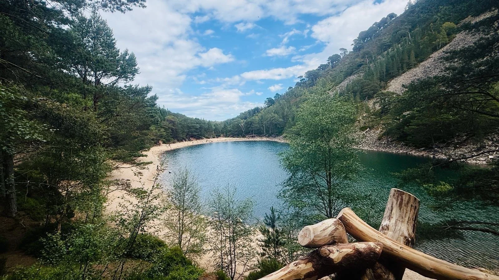 A serene lake surrounded by green trees and forested hills under a partly cloudy sky.