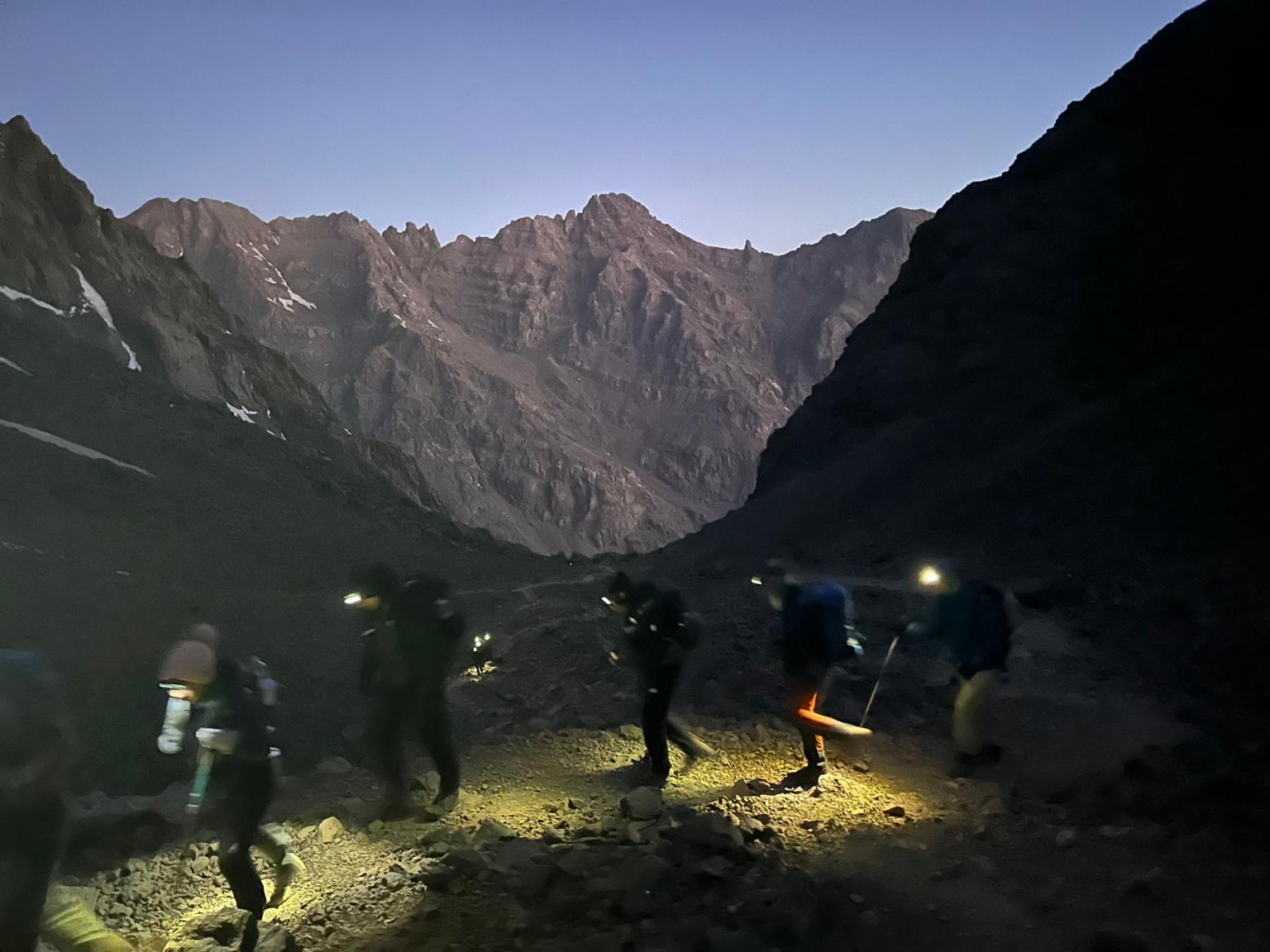 Group of hikers walking through a mountainous terrain at dusk, with a clear sky and rugged mountain peaks in the background, illuminated by their headlamps.