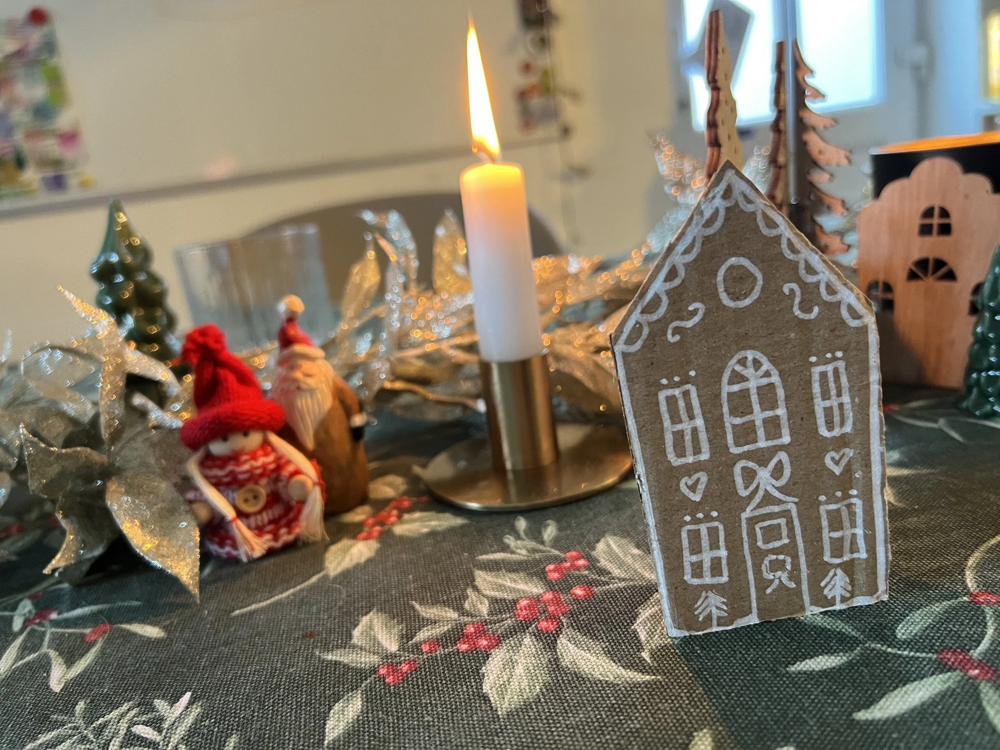 Close-up of a Christmas-themed table centerpiece featuring a lit white candle, decorated wooden house, small Christmas trees, and miniature figurines, set on a festive tablecloth with holly leaves and berries.