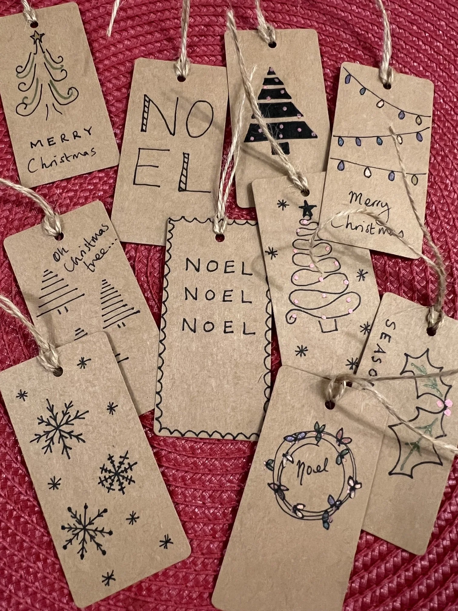 Hand-drawn Christmas-themed gift tags with messages like 'Merry Christmas', 'Noel', and illustrations of Christmas trees, holly, and snowflakes, attached to strings on a Christmas red textured background.