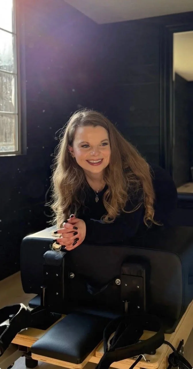 A young woman with long, wavy blonde hair, smiling, sitting at a massage or spa table in a room with dark walls and a window.