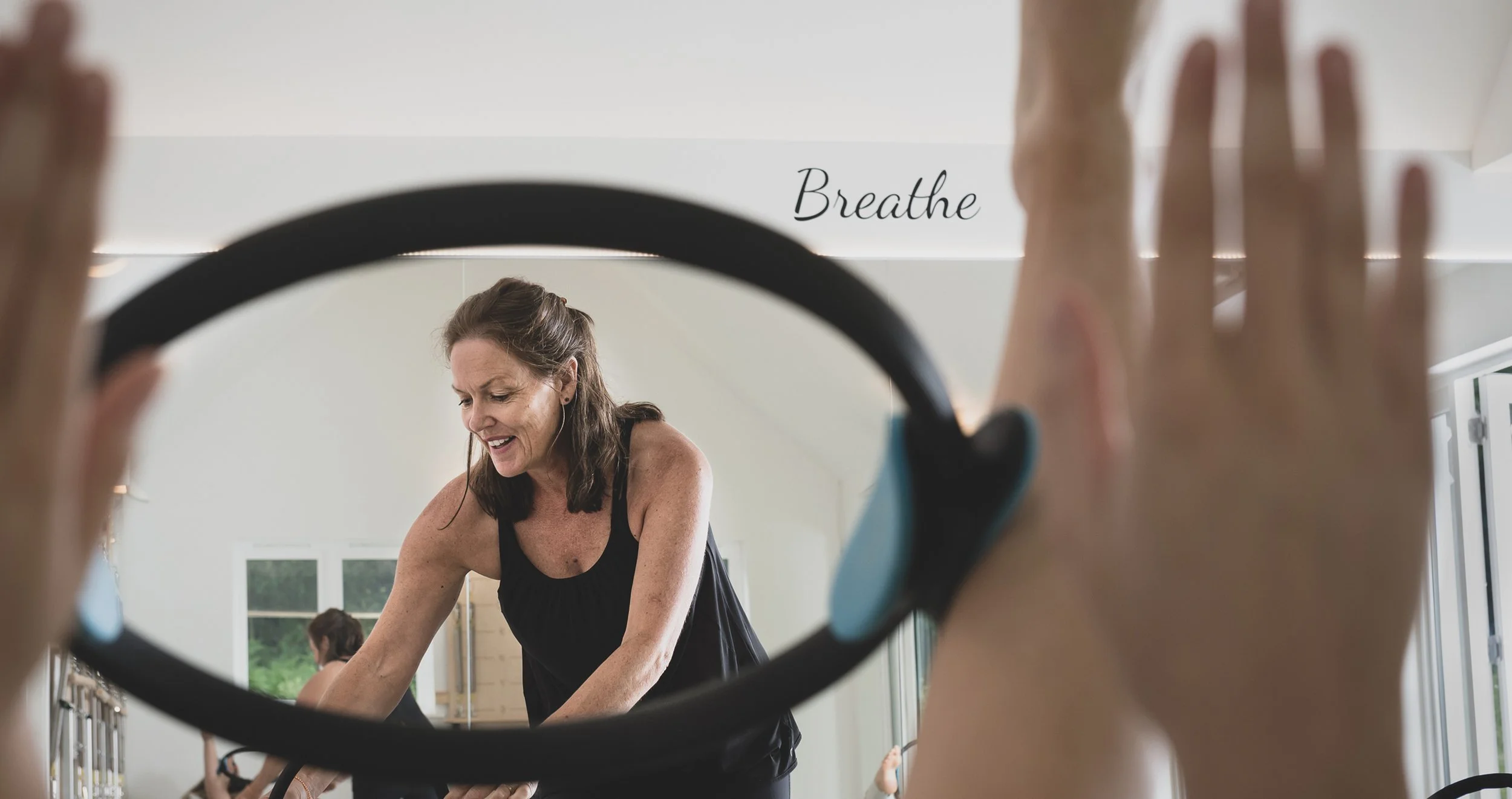 A woman is seen through a fitness hoop, smiling and exercising in a bright gym with the word 'Breathe' written on the wall behind her.