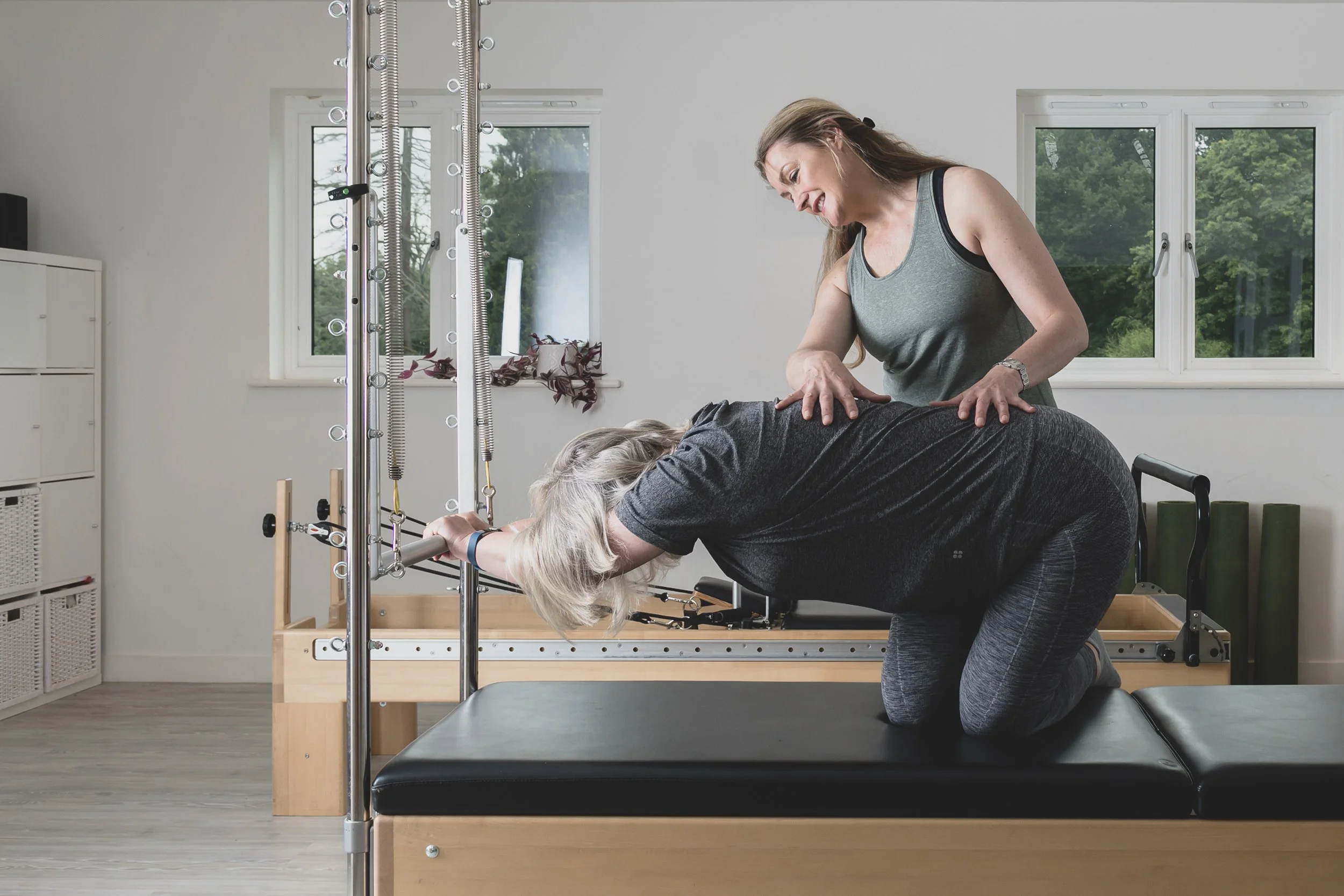 A women in grey exercise clothes performing a physical therapy exercise with her head and arms on a reformer machine, supervised by a therapist in a bright room with large windows and greenery outside.
