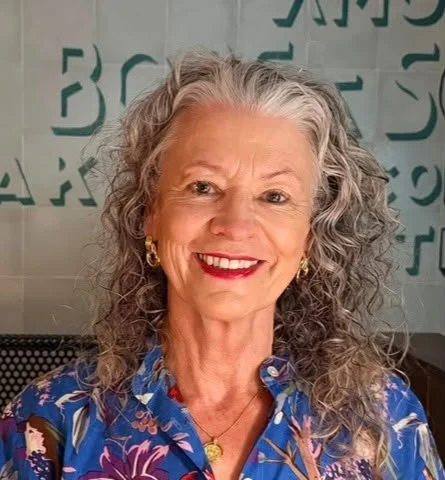 A smiling older woman with curly gray hair, wearing a colorful patterned blouse and gold earrings, sitting indoors with a background of green letters.