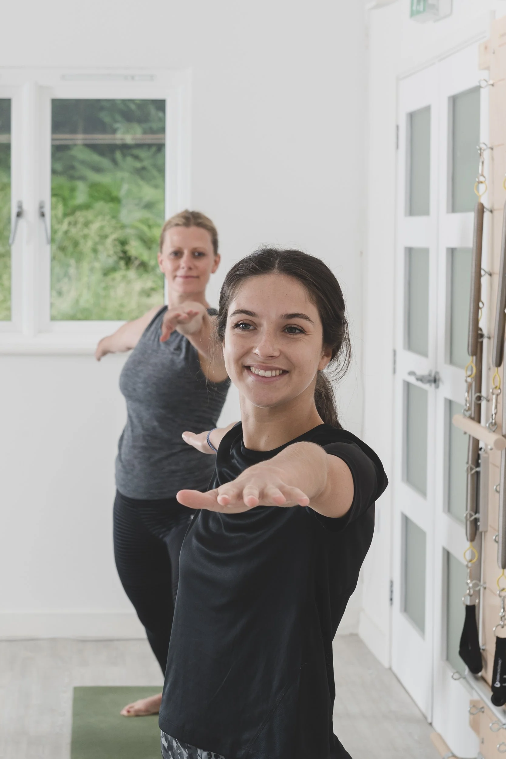Two women doing yoga in a bright room with windows and exercise equipment, both smiling and extending one arm forward with palms facing down.