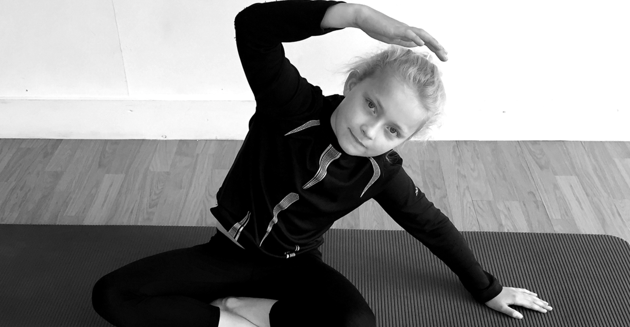 A young girl with light-colored hair and light skin lying on her side on a yoga mat, performing a stretching pose with one arm raised above her head and the other supporting her on the yoga mat.