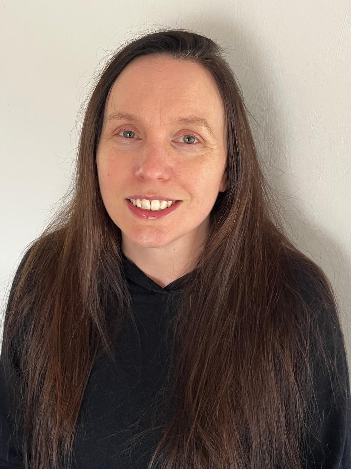 Portrait of a woman with long brown hair wearing a black hoodie, smiling against a plain white wall.