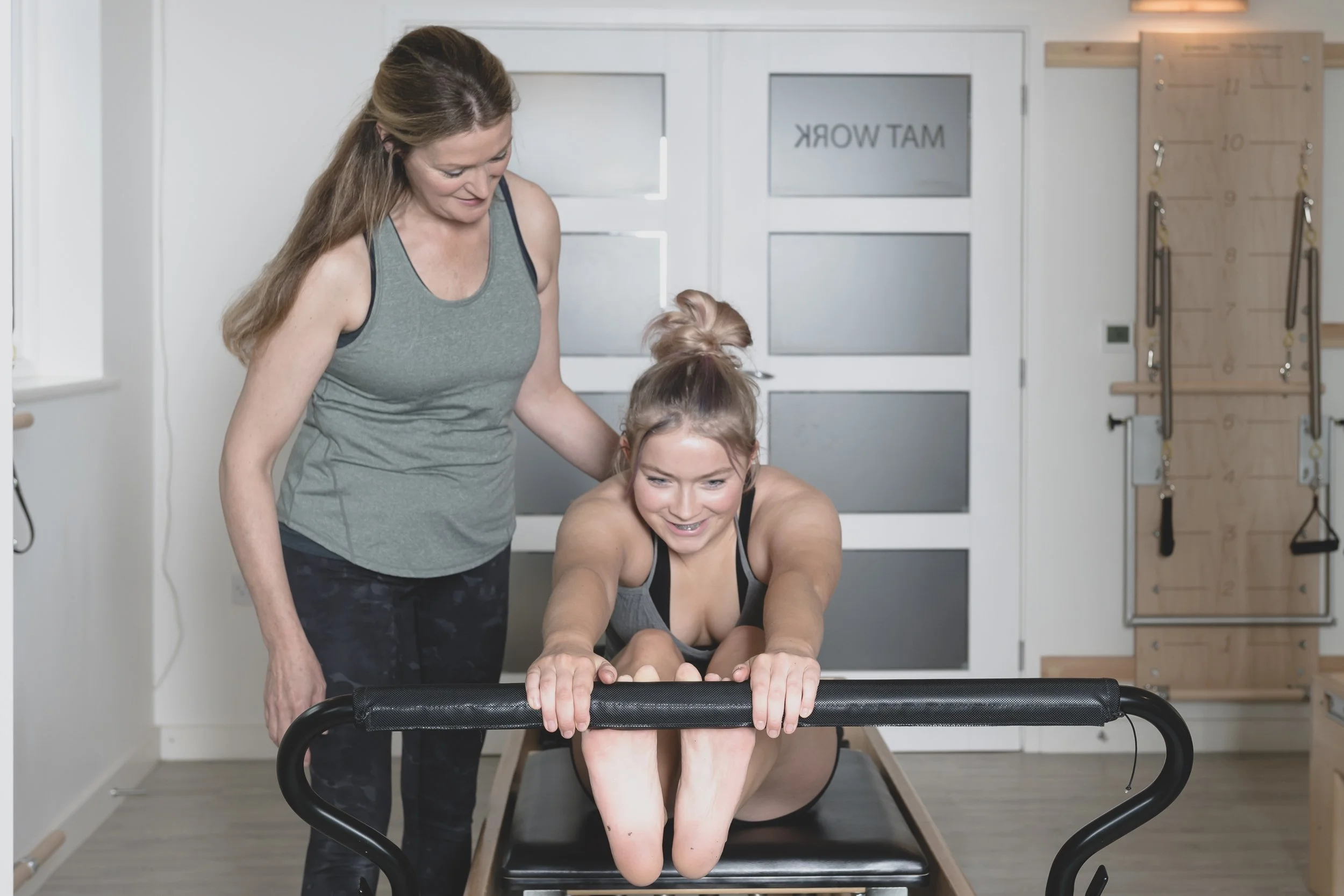 A woman assisting a young woman performing a plank exercise on a Pilates reformer at a fitness studio, with fitness equipment in the background.