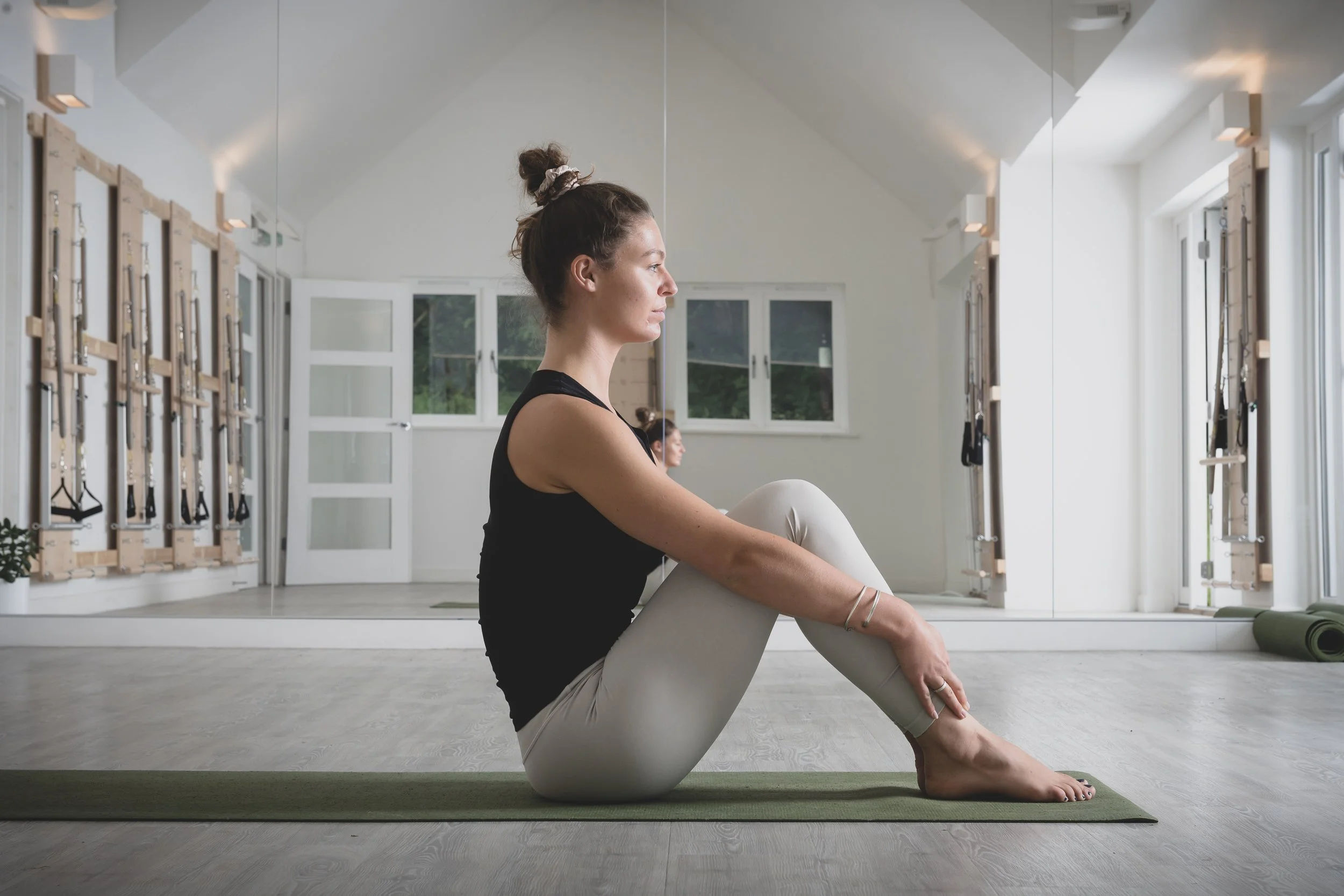 A woman practicing yoga in a bright room, sitting on a yoga mat in a seated forward bend pose with a mirror behind her, workout equipment on the walls, and large windows letting in natural light.
