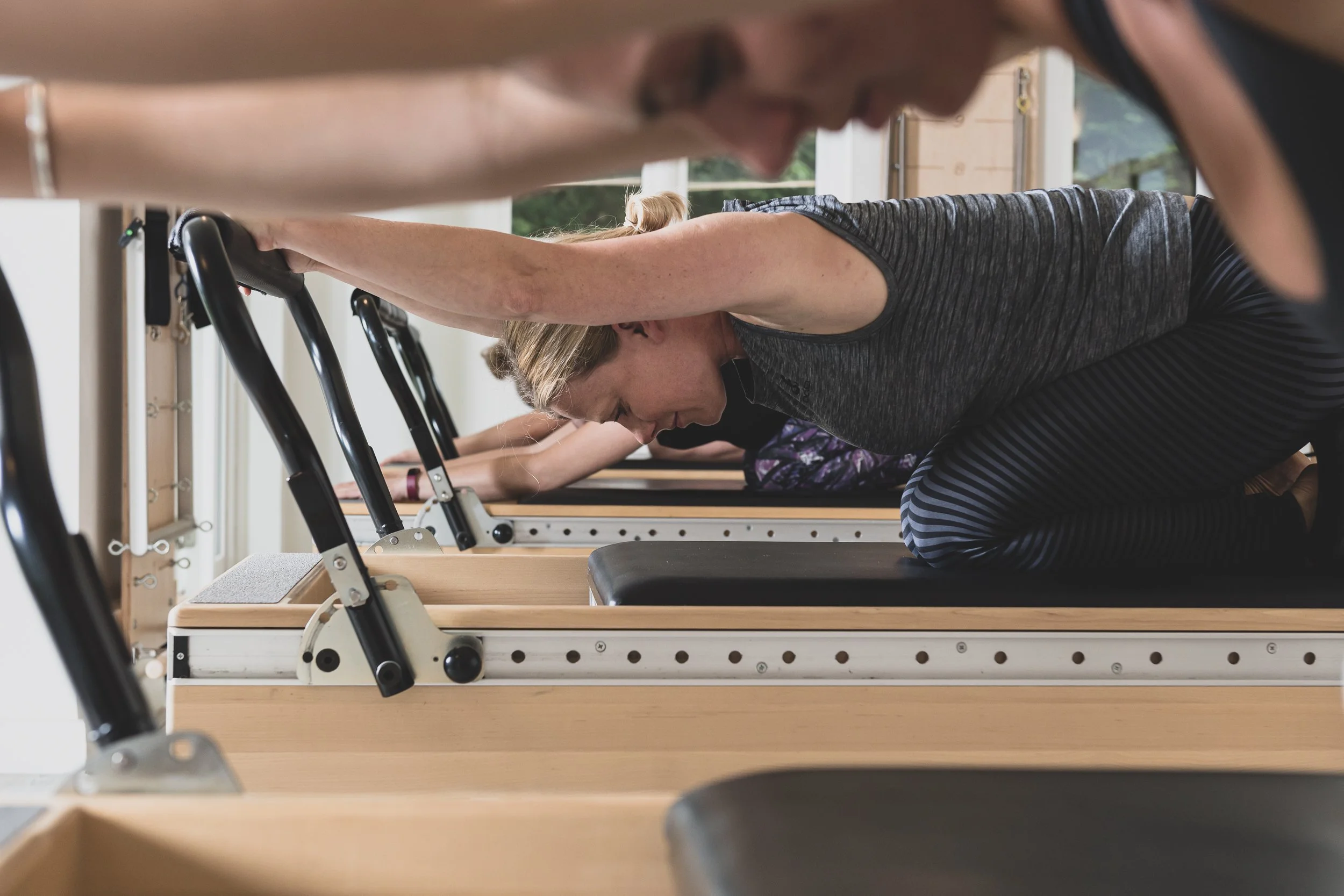 A woman doing Pilates or exercise on a Pilates reformer machine in a bright gym or studio, with other people also exercising in the background.