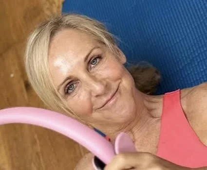 A woman with blonde hair taking a selfie in a room with a wooden floor, holding a pink selfie stick, wearing a pink tank top, and smiling at the camera.