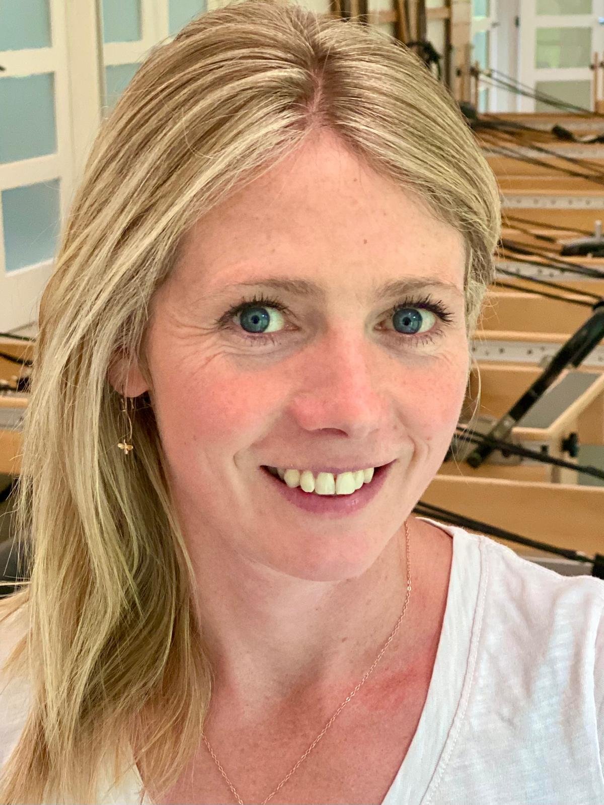 Close-up of a woman with blonde hair, blue eyes, and earrings, smiling in front of a background with wooden chairs and a window.