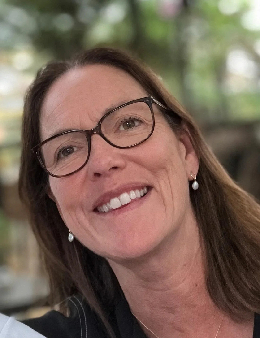 Close-up of a woman with long brown hair, glasses, and pearl earrings, smiling outdoors with blurred greenery in the background.