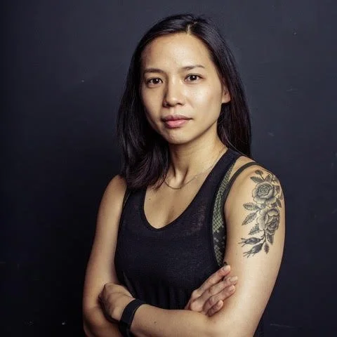 Woman with dark hair and a tattoo on her upper arm, wearing a black sleeveless top, standing against a dark background.