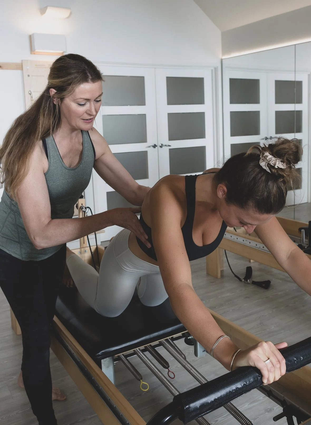 A woman doing a Pilates exercise on a reformer machine under the supervision of an instructor in a studio.
