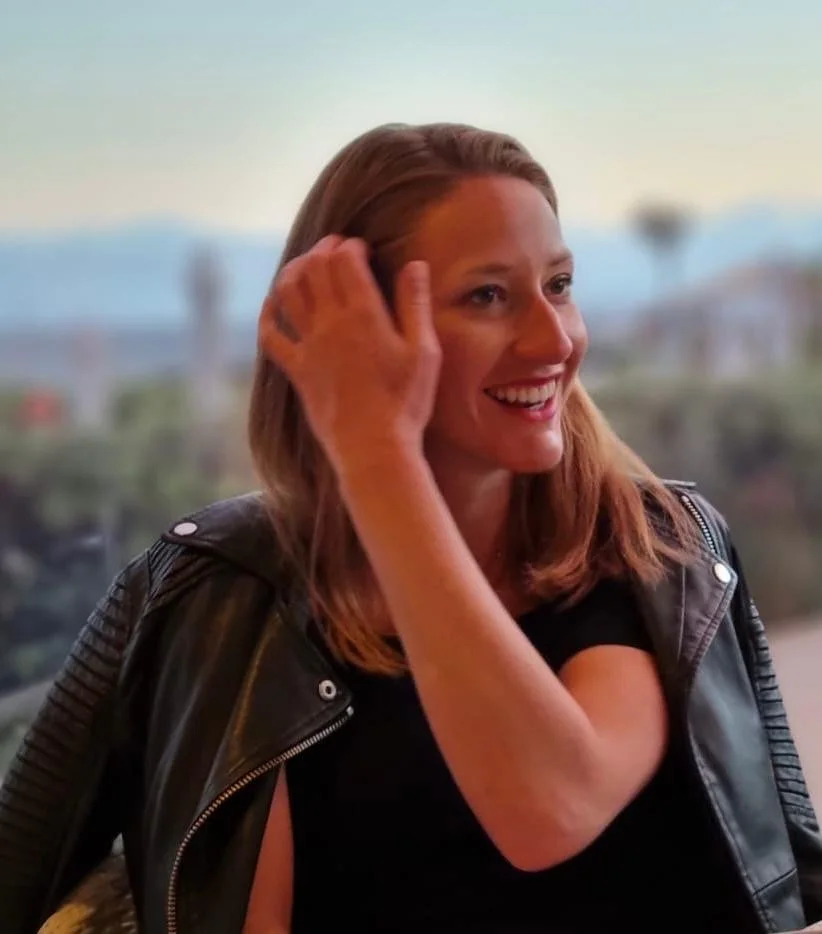 A woman with brown hair smiling, touching her hair, wearing a black shirt and a black leather jacket, with an outdoor cityscape background.