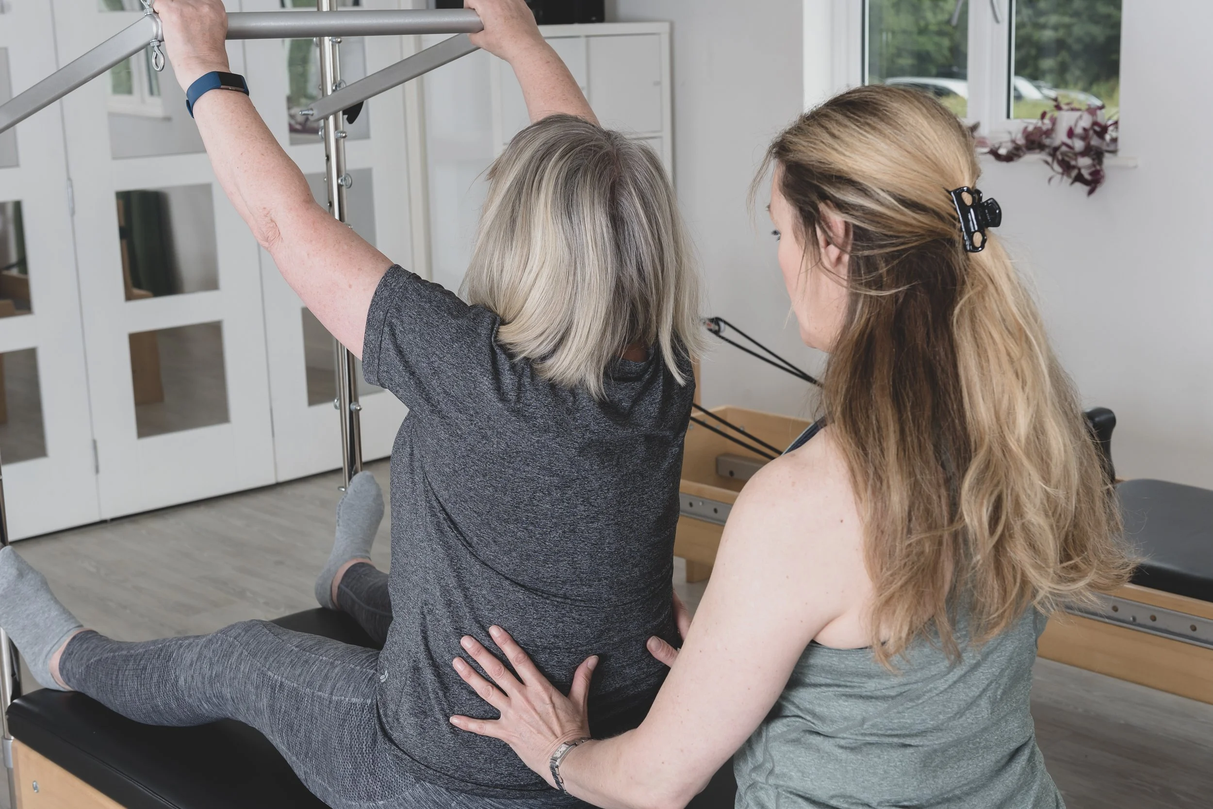 A physical therapist or trainer assisting an elderly woman with a stretching or therapeutic exercise on a reformer machine in a rehabilitation or fitness clinic. The woman is seated on the reformer, extending her arms to hold the bar overhead, while the trainer supports her from behind, offering guidance and support.