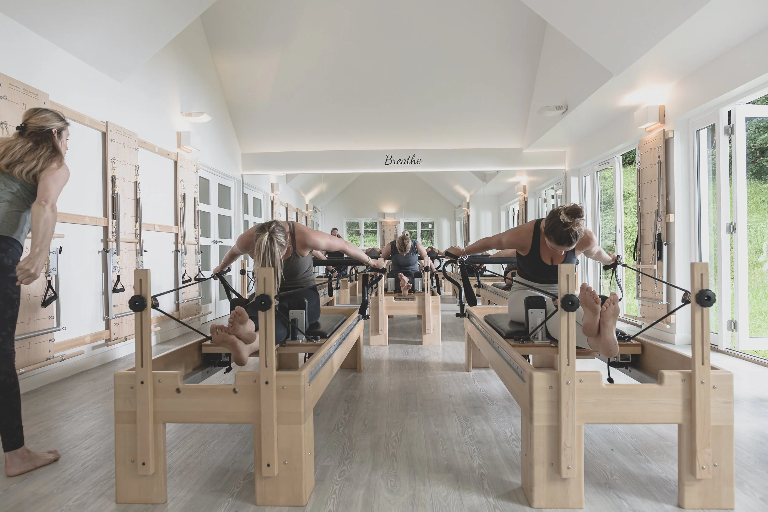 A group of people practicing Pilates using reformer machines in a bright, spacious studio with large windows and a wall sign that says 'Breathe'.
