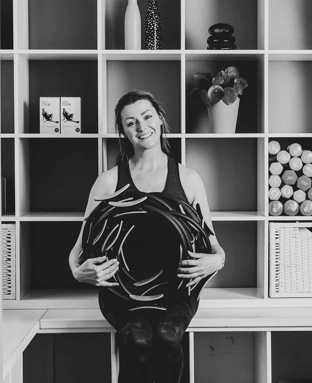 A woman sitting in front of a shelving unit filled with fitness equipment and decorative items, holding a bundle of resistance bands and smiling.