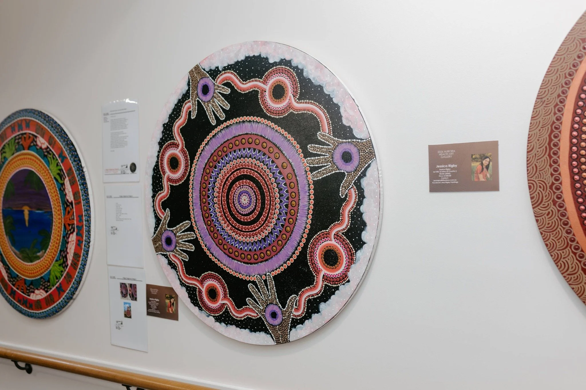 Colorful Aboriginal dot painting with concentric circles, hand shapes, and intricate patterns on a rounded canvas displayed on a gallery wall.