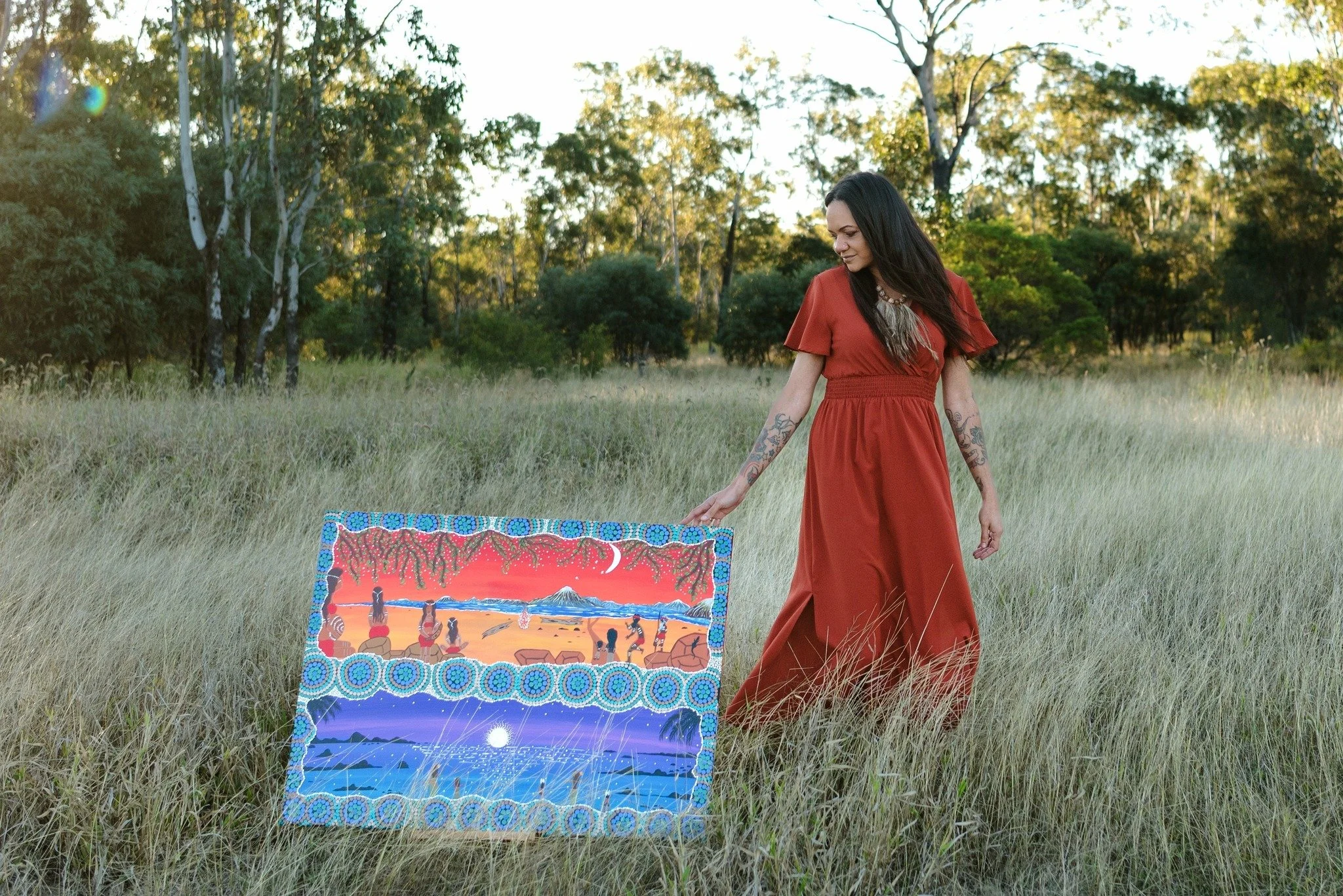 A woman in a red dress standing in a grassy field near trees, looking at a colorful painting on the ground depicting a sunset or sunrise over mountains with figures in the foreground.