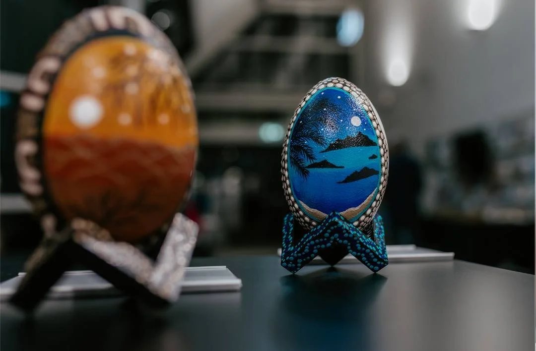 Decorative oval ornaments, each with painted landscape scenes, one featuring a sunset or sunrise over mountains, and the other depicting a moonlit lake scene with trees and mountains, displayed on stands.