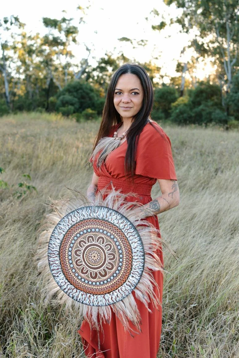 A woman with long dark hair standing in a field of tall grass, holding a decorated circular shield with a fur trim. She is wearing a rust-colored dress and a necklace, with a tattoo visible on her arm, and a background of trees at sunset.