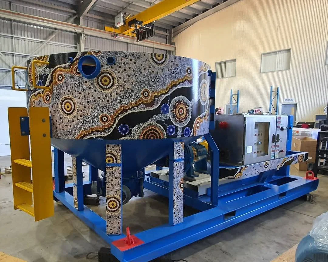 Large industrial machine with Aboriginal art design in black, yellow, white, and blue, situated in a warehouse with metal walls and a yellow overhead crane.