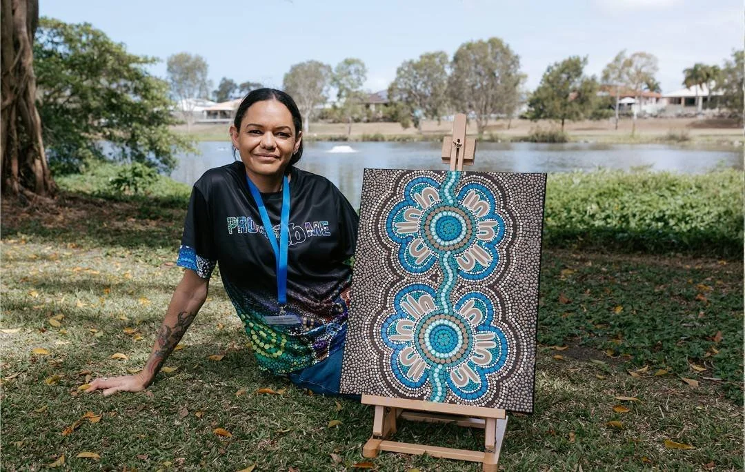 A woman sitting on the grass outdoors near a lake with a diamond painting artwork on an easel next to her.