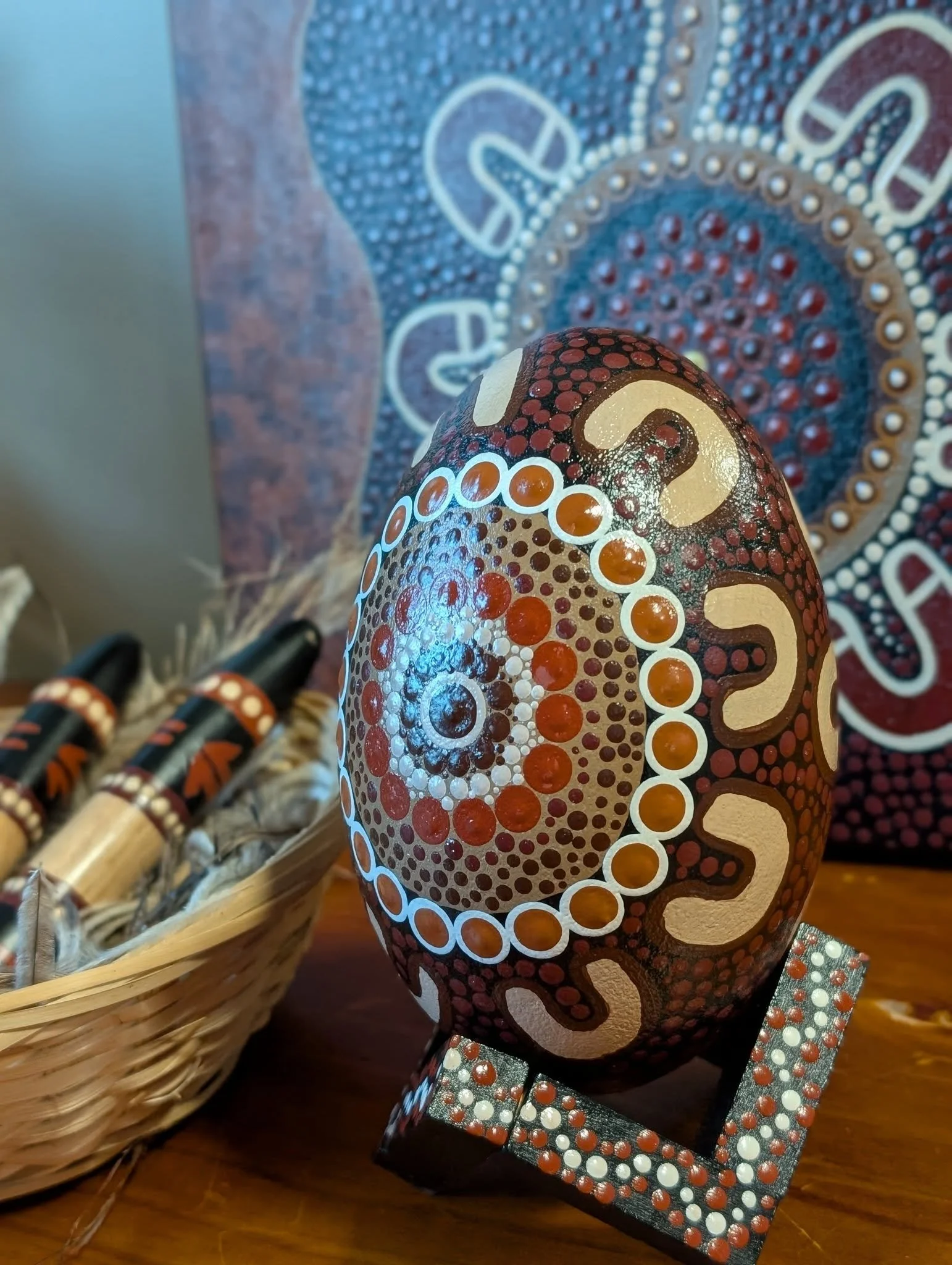 Decorative Aboriginal-style painted rock with dot patterns, resting on a table next to a woven basket with other painted items inside.