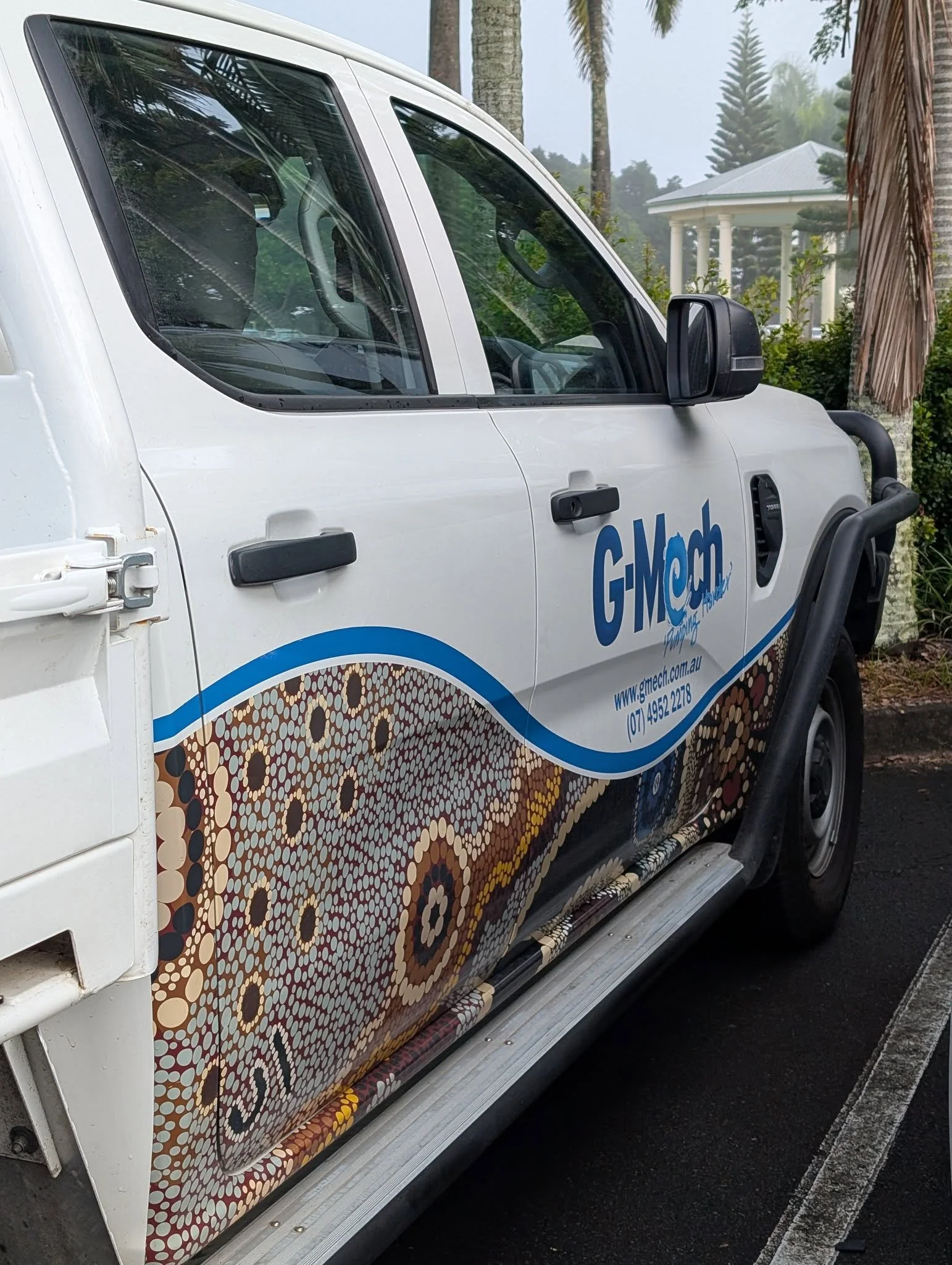 A white vehicle with colorful dot patterns and the logo 'G-MECH' on the side, parked next to a paved road with a curb. The background features palm trees and a white pavilion with columns, in a lush, green environment.