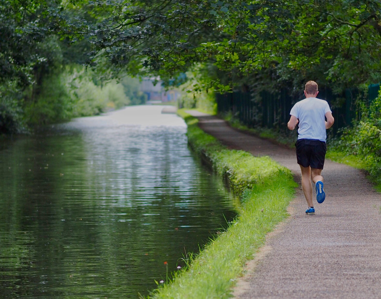 A man jogging on a path beside a canal or river, with green trees overhead and lush grass along the water.