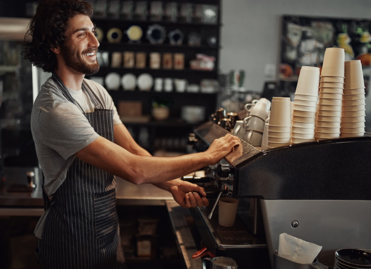 A smiling man working behind the counter at a coffee shop, preparing to operate an espresso machine with stacked paper cups nearby.
