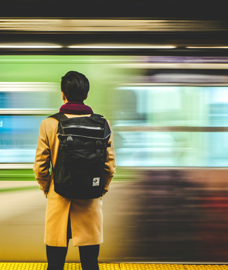 Person with black hair wearing a tan coat and maroon scarf, carrying a black backpack, standing on a yellow tactile paving, as a green and purple train speeds by in a subway station.
