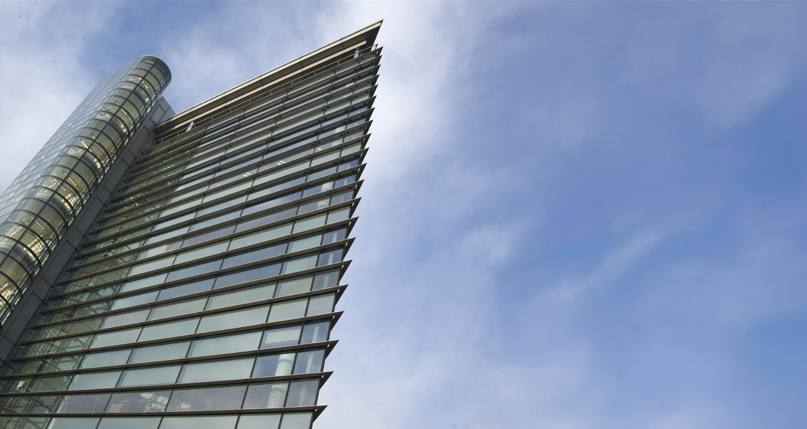 A modern high-rise building with glass windows and metal framework, viewed from below against a blue sky with a few clouds.