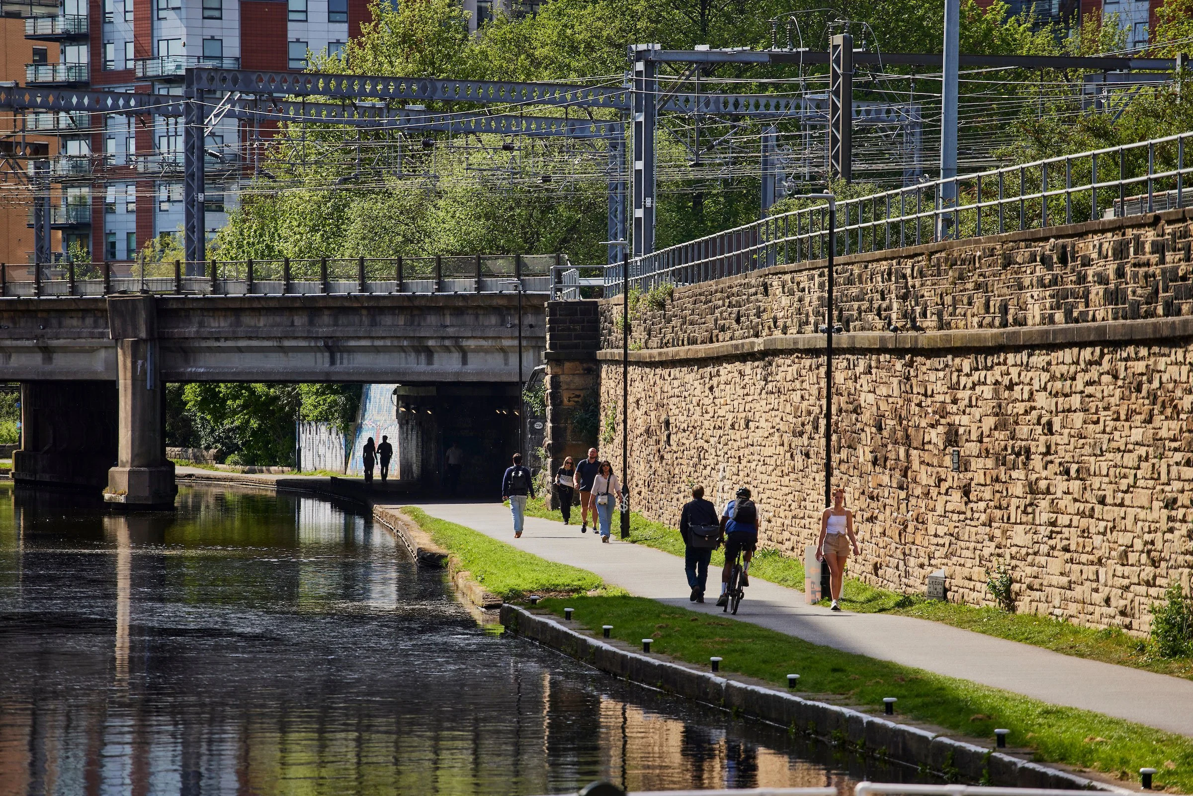People walking and cycling along a riverwalk path beneath a bridge in an urban area, with a stone wall and apartment buildings in the background.