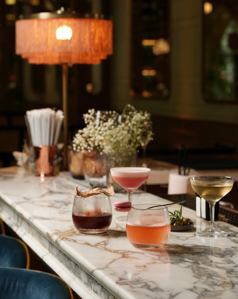 A marble bar table with four cocktails, a flower arrangement, and a copper lamp in a dimly lit bar or restaurant.