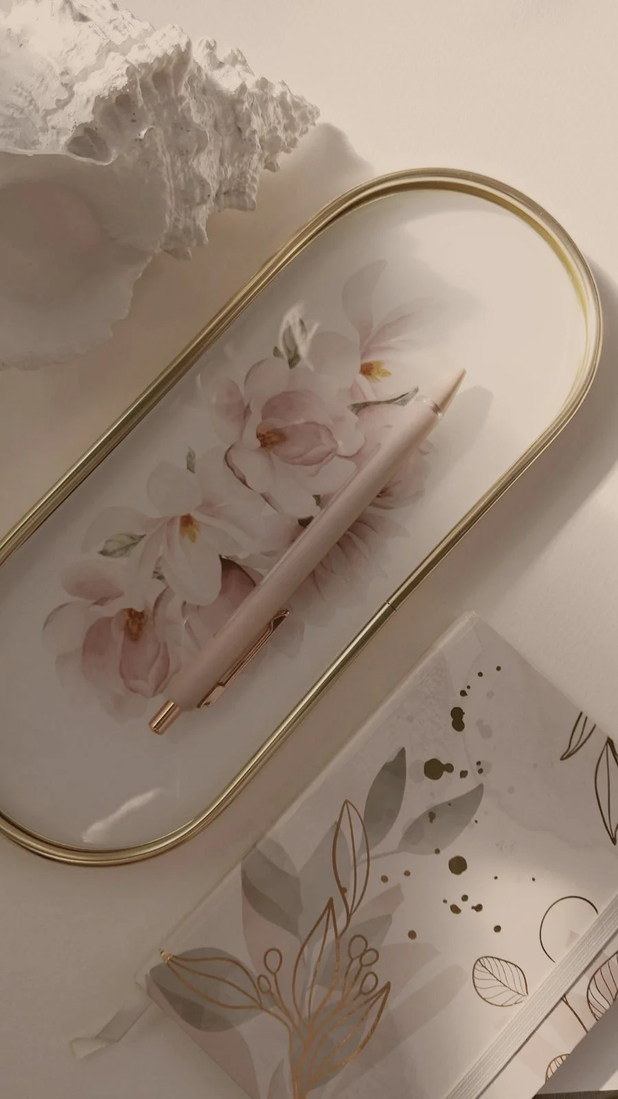 Decorative tray with pink and white flowers design, a white pen, a white decorative object with a textured surface, and a white box with gold leaf and dot patterns.