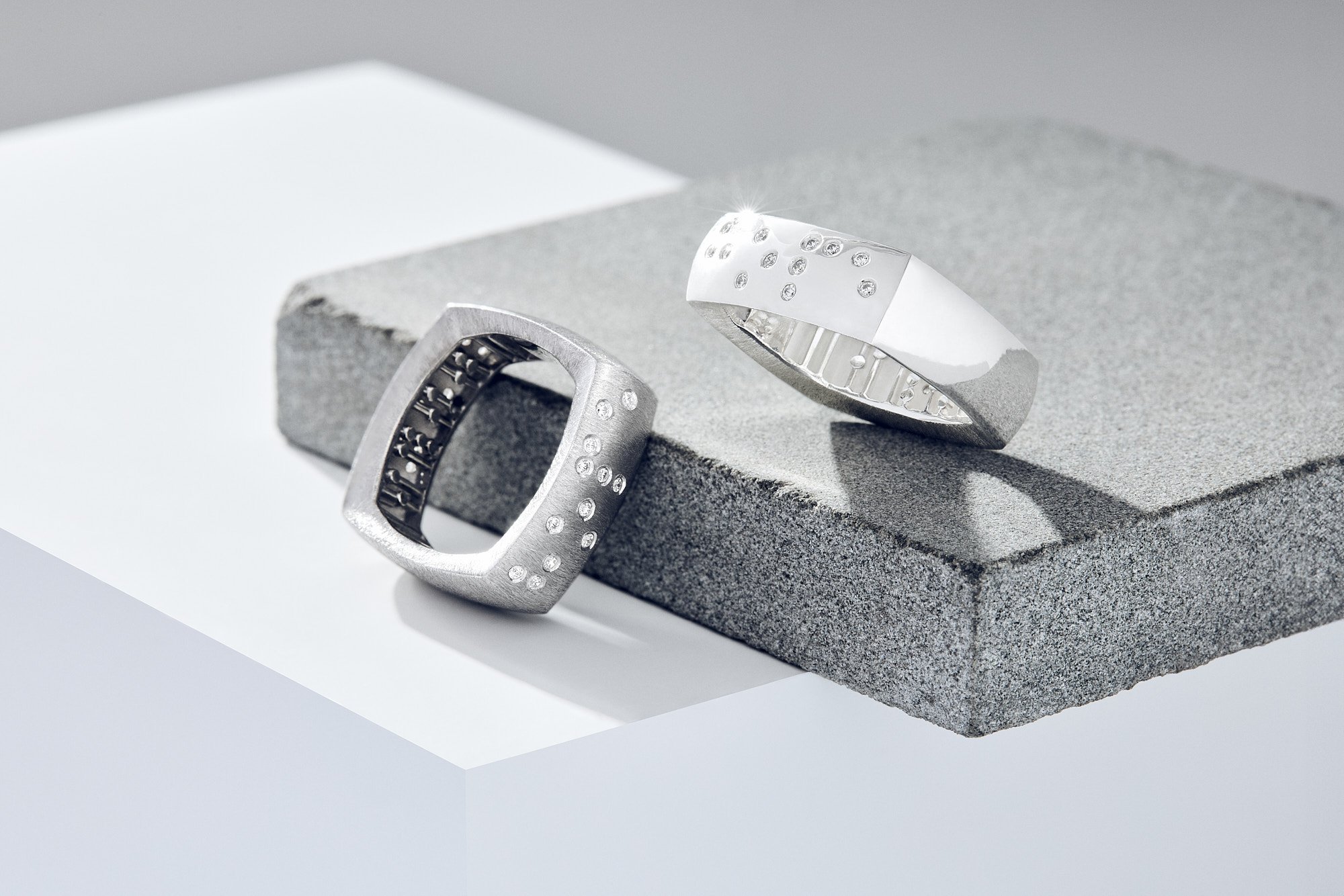 Two silver rings with embedded crystals, one with a large square face and the other a rounded band, displayed on a gray and white textured surface.