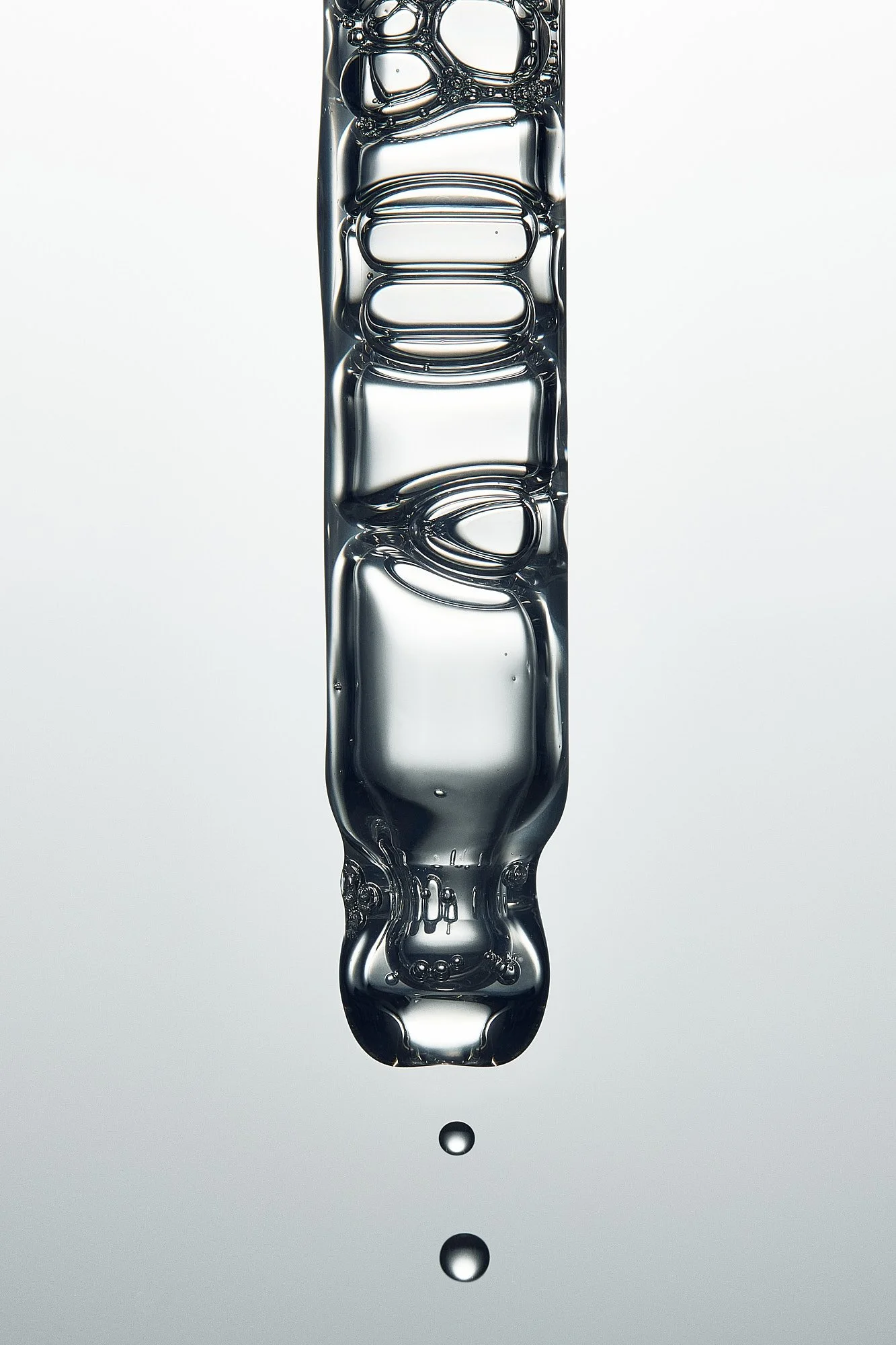 Close-up of a drip of water falling from a crystal-clear, elongated water droplet with a reflective surface, against a plain, light gray background.