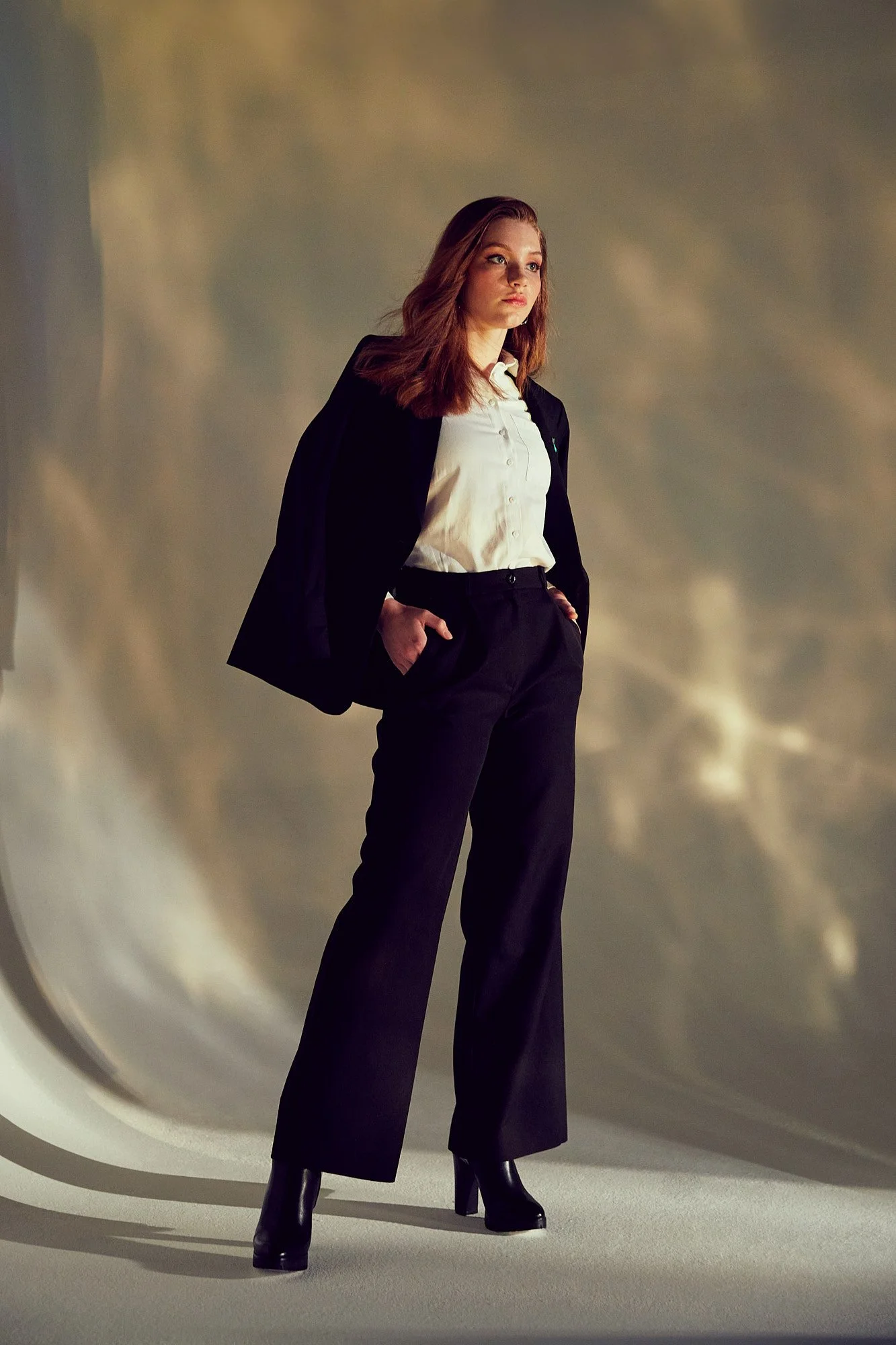 A woman with red hair wearing a white shirt, black blazer, and wide-leg black pants, standing with hands in pockets inside a reflective, metallic tunnel.