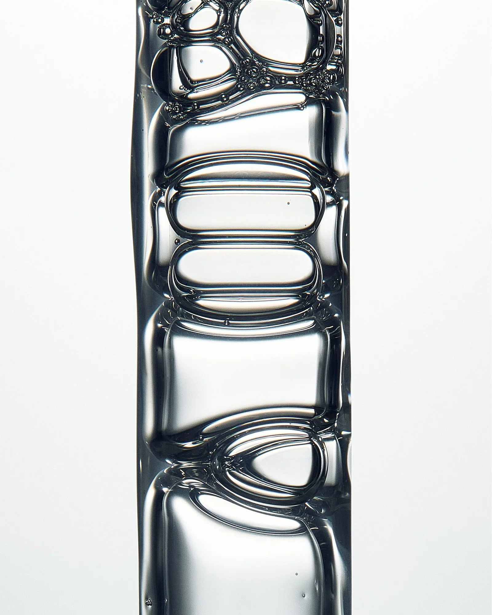 Close-up of a drip of water falling from a crystal-clear, elongated water droplet with a reflective surface, against a plain, light gray background.