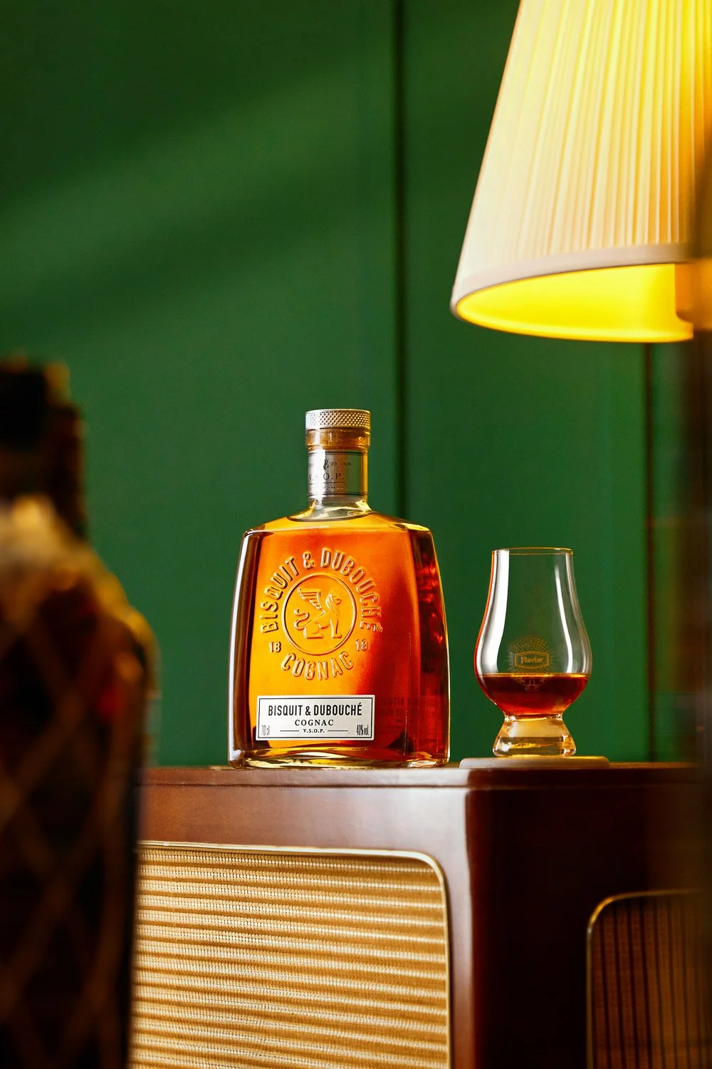 A bottle of Bisquit & Dubouché Cognac with a glass of cognac on a wooden surface, green wall background, and a yellow lampshade.