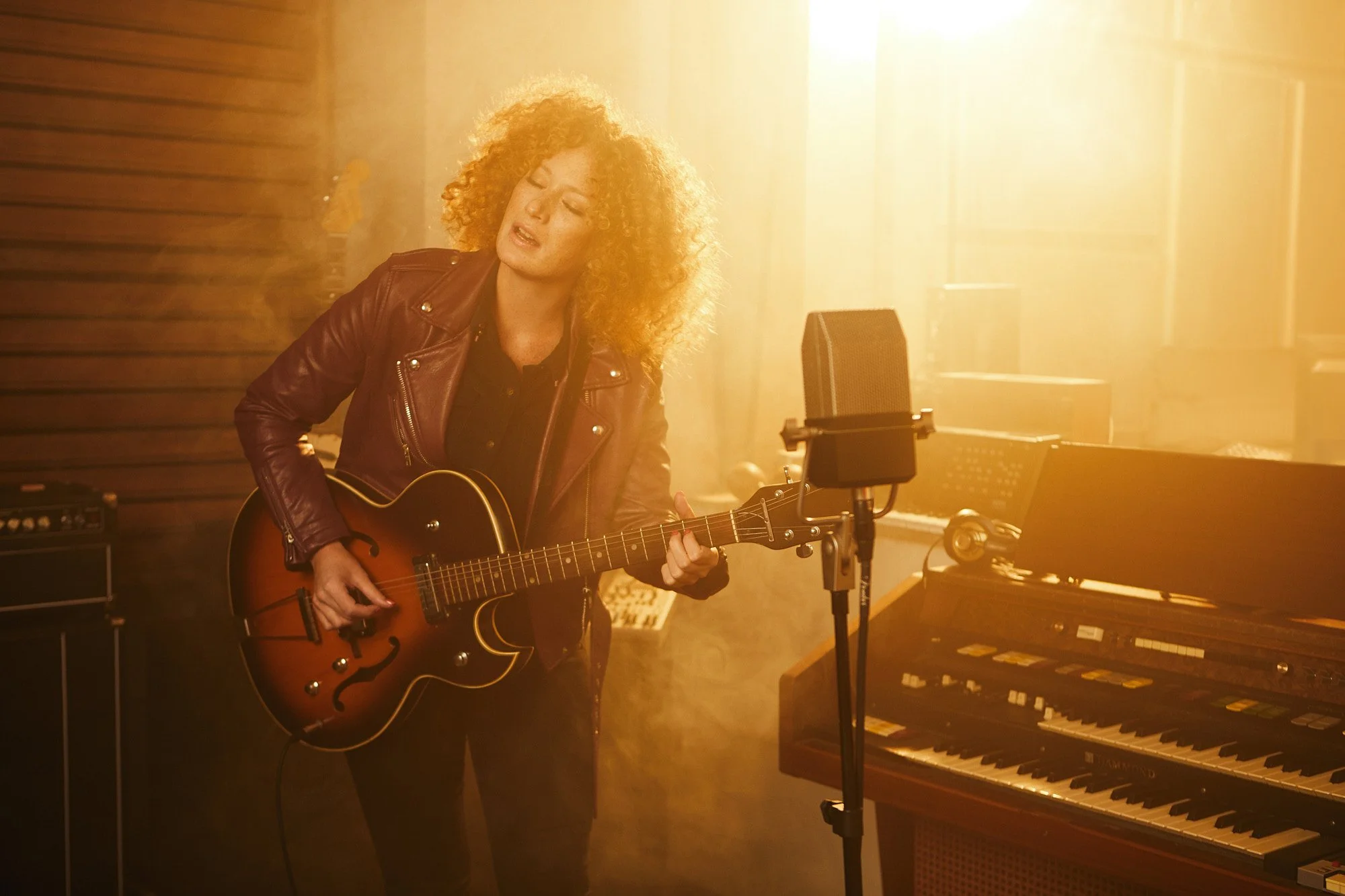A Marina Martensson with curly red hair playing an acoustic guitar in a music studio, surrounded by recording equipment, with sunlight streaming through a window.