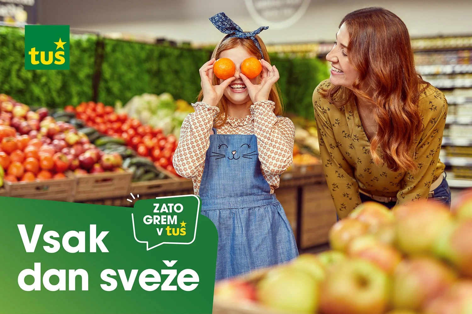 A young girl with red hair, wearing a blue apron with a cat face, holding two oranges over her eyes and smiling. An adult woman with long red hair, wearing a yellow blouse with small black patterns, is smiling and looking at her in a grocery store pr