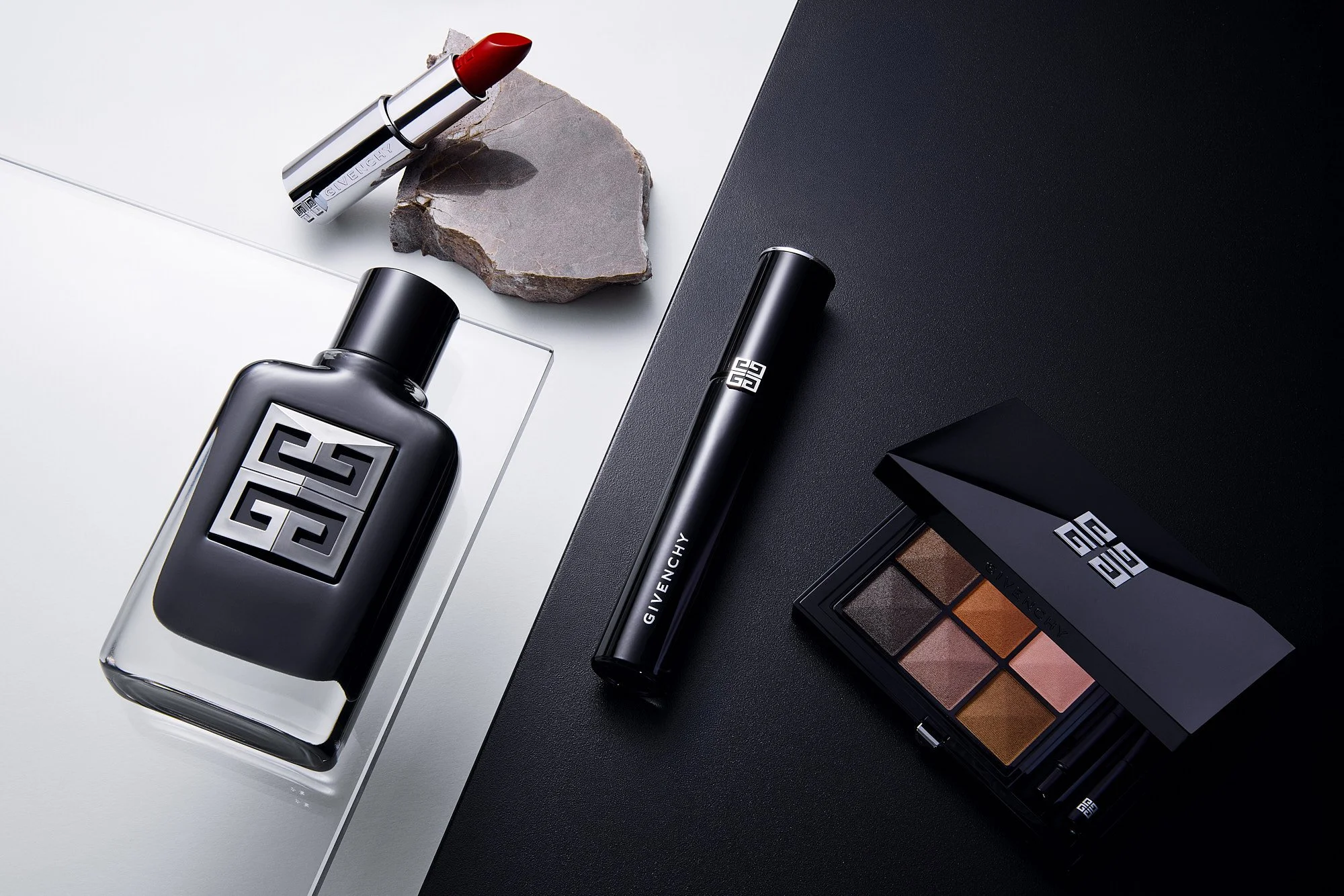 A flat lay of Givenchy beauty products, including a lipstick, a perfume bottle, a eyeliner, eyeshadow palette, and a Paco Rabanne perfume bottle, arranged on a black and white surface with a gray stone.