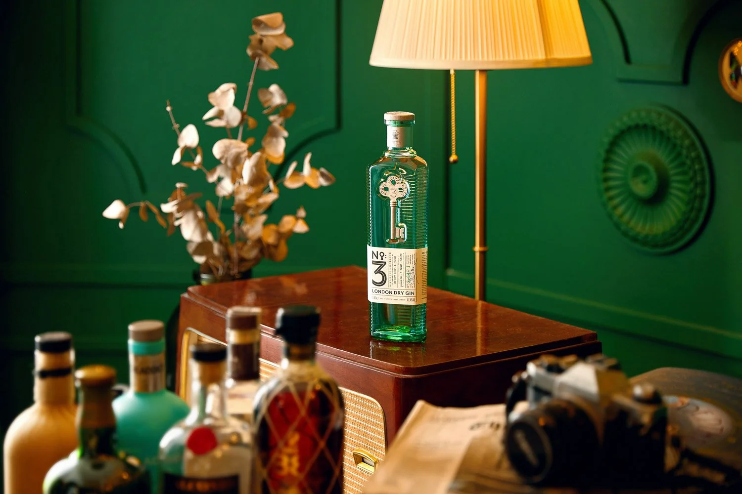 A green bottle of London Dry Gin with a label, placed on a wooden table, with a dried plant in the background, surrounded by various other bottles, a lamp, and a camera, in a decoratively paneled green room.