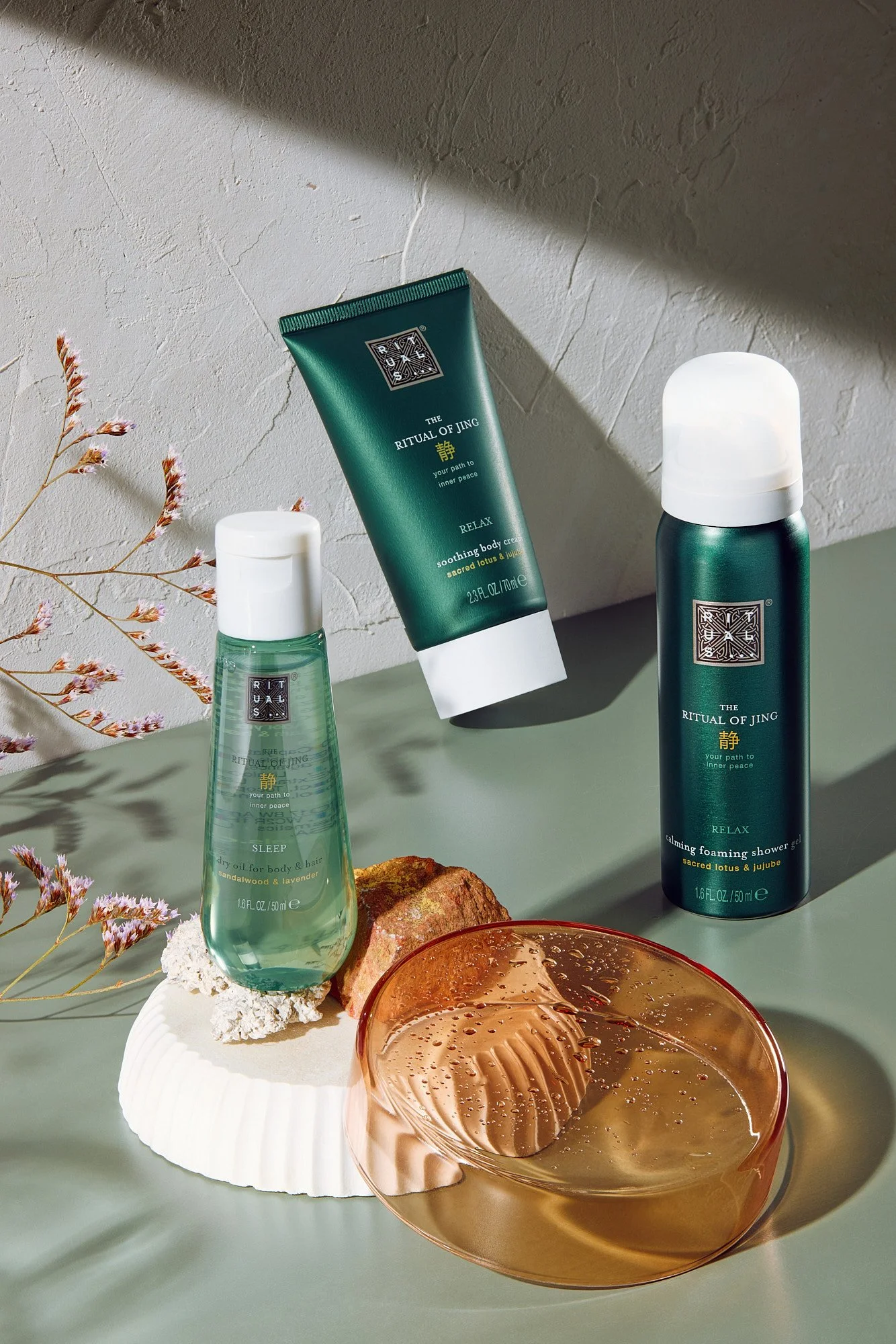 A collection of skincare products, including a calming foaming shower gel, a soothing body cream, and a sleep oil, arranged on a pale green surface with dried flowers and a decorative shell.